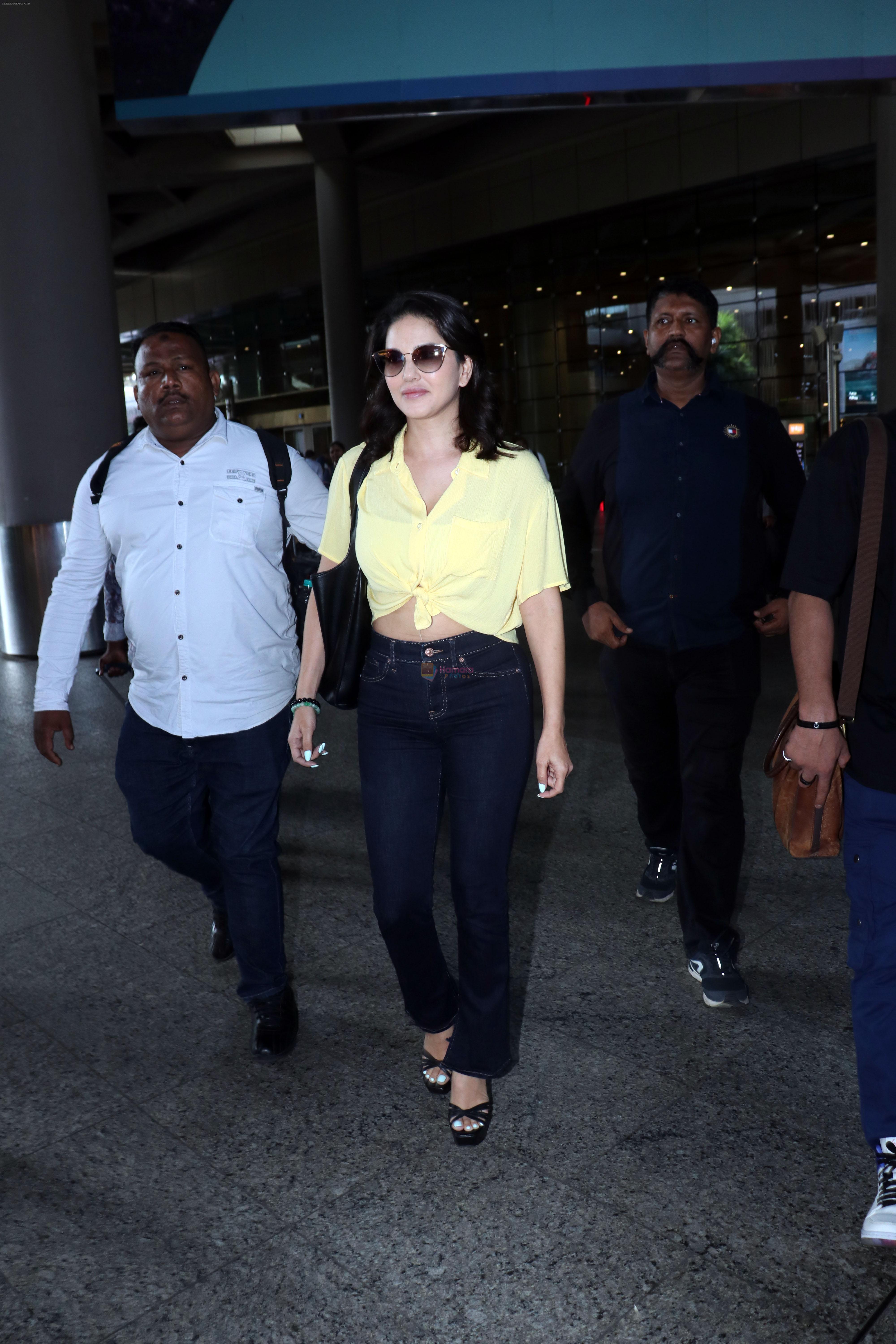 Sunny Leone is dressed in a yellow shirt blue jeans sunglasses and black high heels