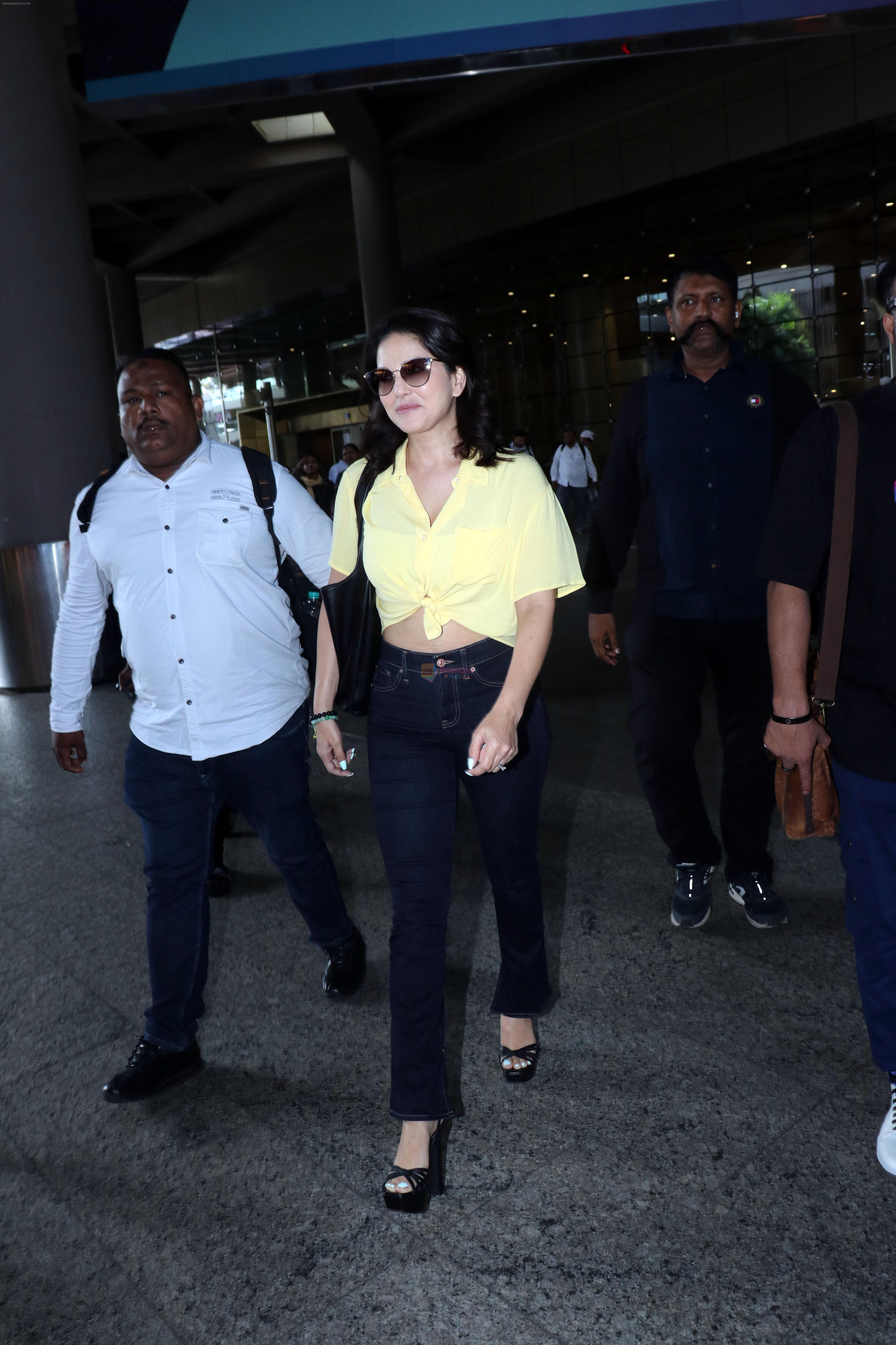 Sunny Leone is dressed in a yellow shirt blue jeans sunglasses and black high heels